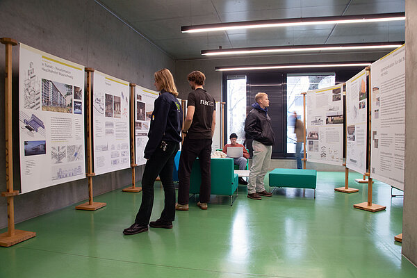 Im Foyer in der Hochschulbibliothek stehen Ausstellungstafeln, Personen besichtigen diese