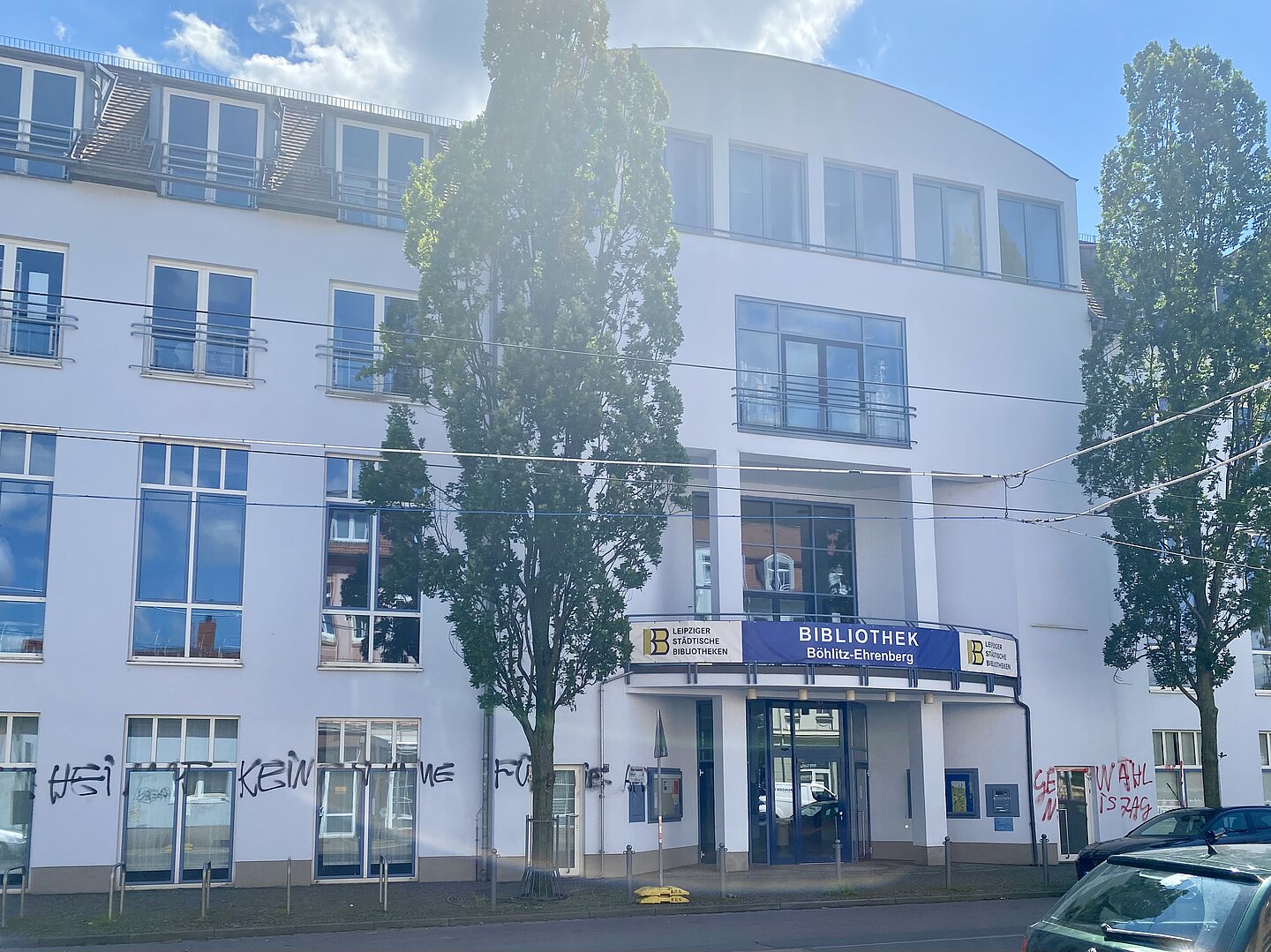 Das Bild zeigt ein modernes, mehrstöckiges Gebäude mit großen Fenstern und einer weißen Fassade. Auf dem Gebäude befindet sich ein Schild mit der Aufschrift „BIBLIOTHEK BÖHLITZ-EHRENBERG“. Das Schild enthält auch das Logo und den Namen „Leipziger Städtische Bibliotheken“. Vor dem Gebäude stehen zwei hohe Bäume. An der unteren Gebäudefassade ist Graffiti zu sehen. Das Gebäude hat mehrere Etagen mit Balkonen im obersten Stockwerk und einen Haupteingang mit Glastüren. Vor dem Gebäude sind Stromleitungen sichtbar.