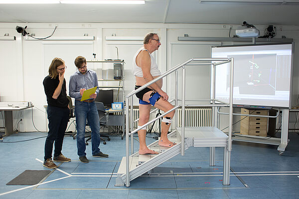 Die Forscher Felix Weiske und Max Böhme stehen neben einem Mann, der am Knie Tracking-Punkte trägt und im Labor eine Treppe hinaufsteigt. 