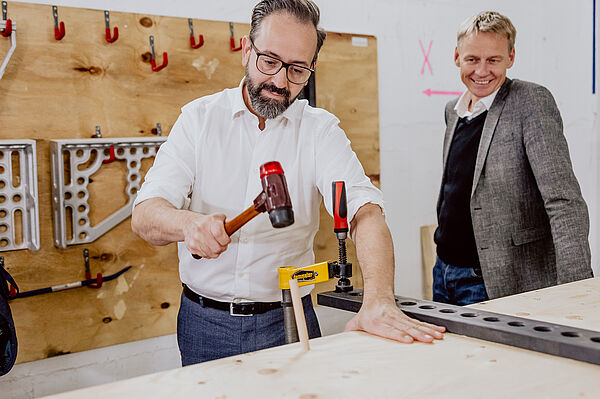 Sebastian Gemkow hämmert eigenhändig einen Holzdübel in eine Holzwandkonstruktion, neben ihm steht Prof. Alexander Stahr