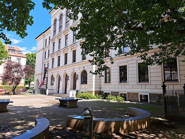 Ein großes mehrstöckiges Altbaugebäude mit weißer Fassade und braunen Fensterrahmen und 3 braunen Türen. Die Fenster im obersten Stockwerk sind sehr groß und bogenförmig. Vor dem Gebäude befindet sich ein gepflasterter Vorplatz, auf dem steinerne Tischtennisplatten stehen. Ein Baum ragt rechts ins Bild.