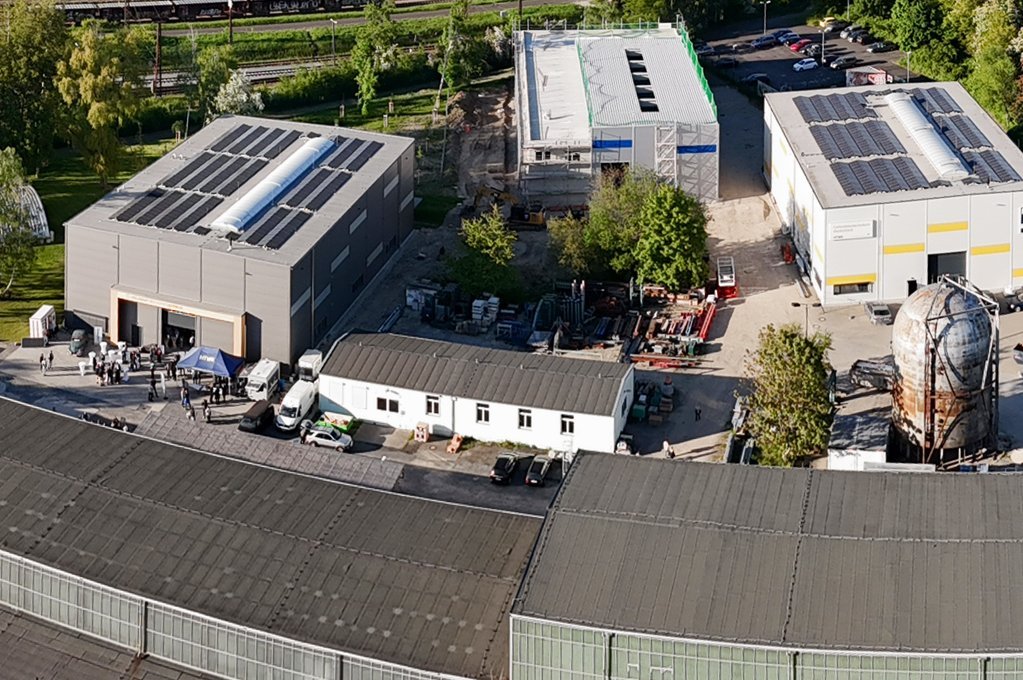 Drohnenaufnahme vom Campus mit mehrere großen Hallen in der Nähe von Wald und Schienen.