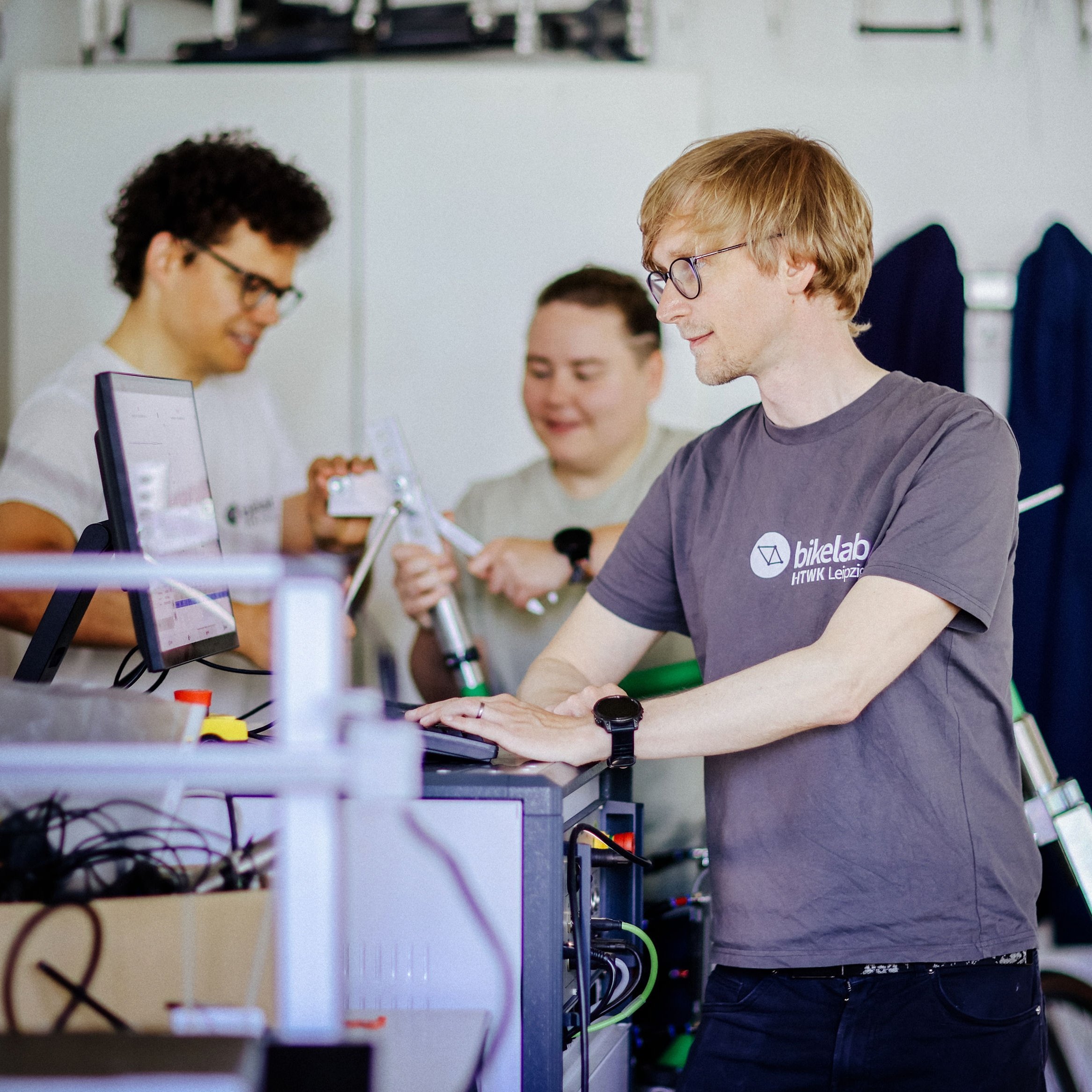 Drei Personen im Bikelab der HTWK Leipzig, eine Person tippt an einem Computer, eine andere hält ein technisches Fahrradbauteil