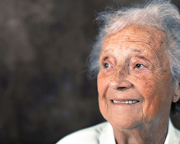 Es ist ein seitliches Porträtfoto einer alten Dame zu sehen. Sie lächelt.
