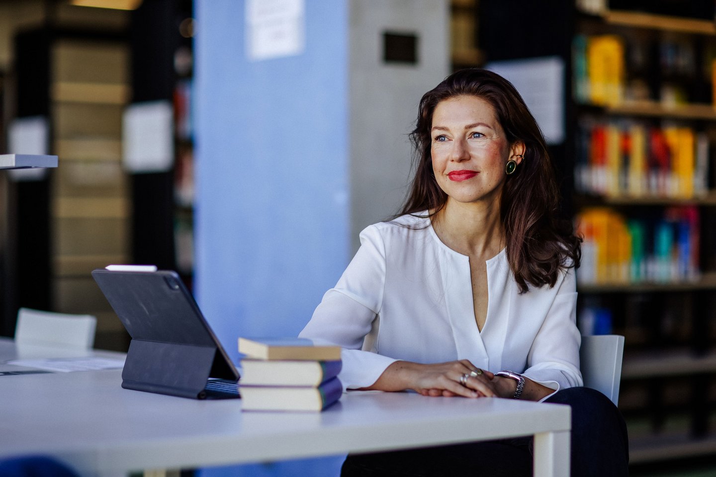 Frau mit langen Haaren sitzt an einem Tisch mit Büchern und Laptop, lächelt in die Kamera.
