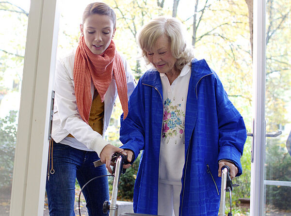 Eine pflegende Person hilft einer pflegebedürftigen Person beim Gang aus dem Garten in einen Innenraum. 