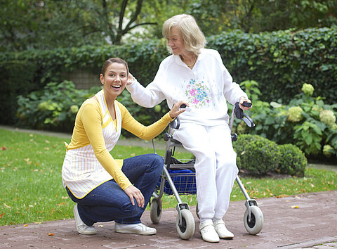 Enkelin mit Oma im Park Oma sitzt auf Rollator im Park. Daneben hockt ihre Enkelin, welche die Oma pflegt.