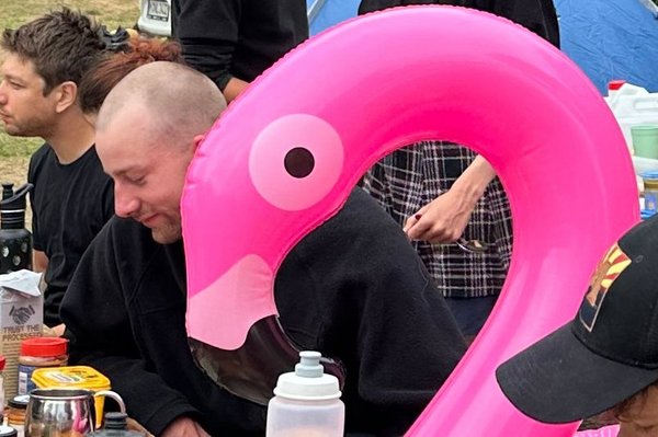Mehrere Personen an einem üppigen Frühstückstisch unter freiem Himmel, zwischen ihnen sitzt ein aufblasbarer Flamingo-Schwimmreif auf der Bank.
