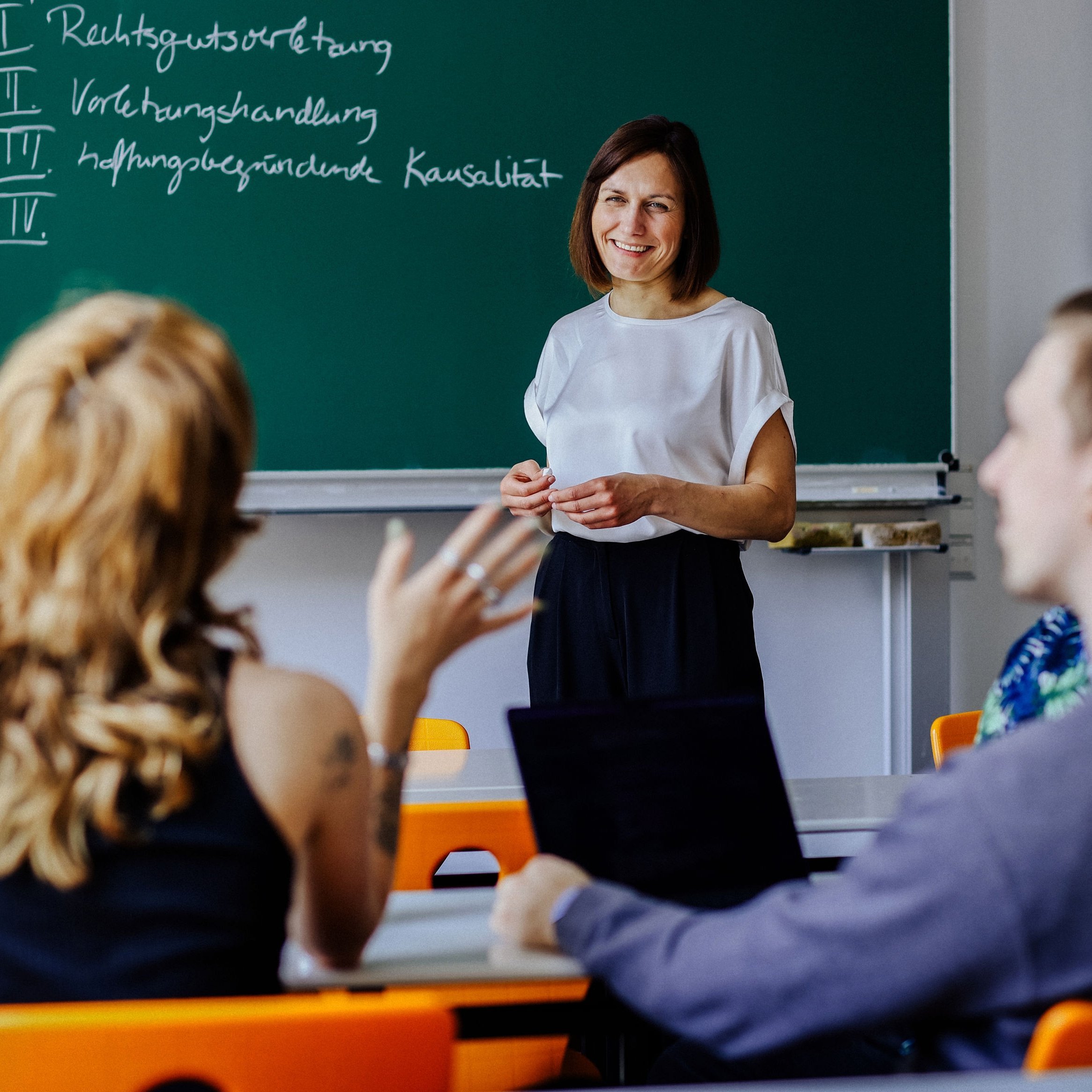 Dozentin steht vor einer Tafel, Studierende diskutieren im Seminarraum.