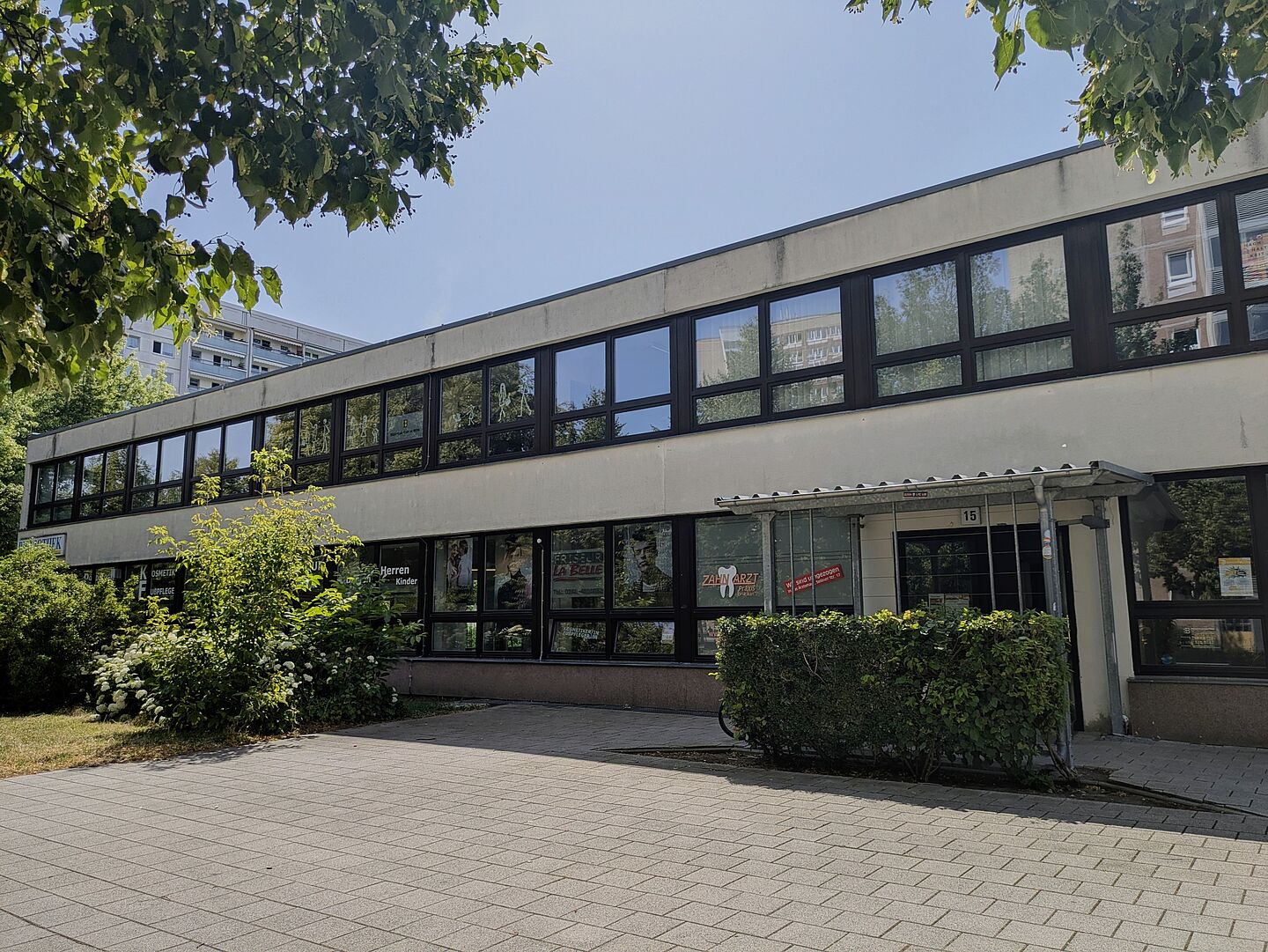 Die Stadtteilbibliothek Grünau-Mitte liegt in einem zweistöckiges Gebäude mit vielen Fenstern. Die Fassade ist beige und die Fensterrahmen sind dunkelbraun. Vor dem Gebäude befindet sich eine gepflasterte Fläche und eine kleine grüne Hecke, die den Eingang säumt. Links neben dem Eingang ist die Werbung einer Zahnarztpraxis sichtbar. Ein Baum und einige Sträucher sind im Vordergrund zu sehen, die etwas Schatten spenden. Im Hintergrund sind weitere Wohngebäude erkennbar.