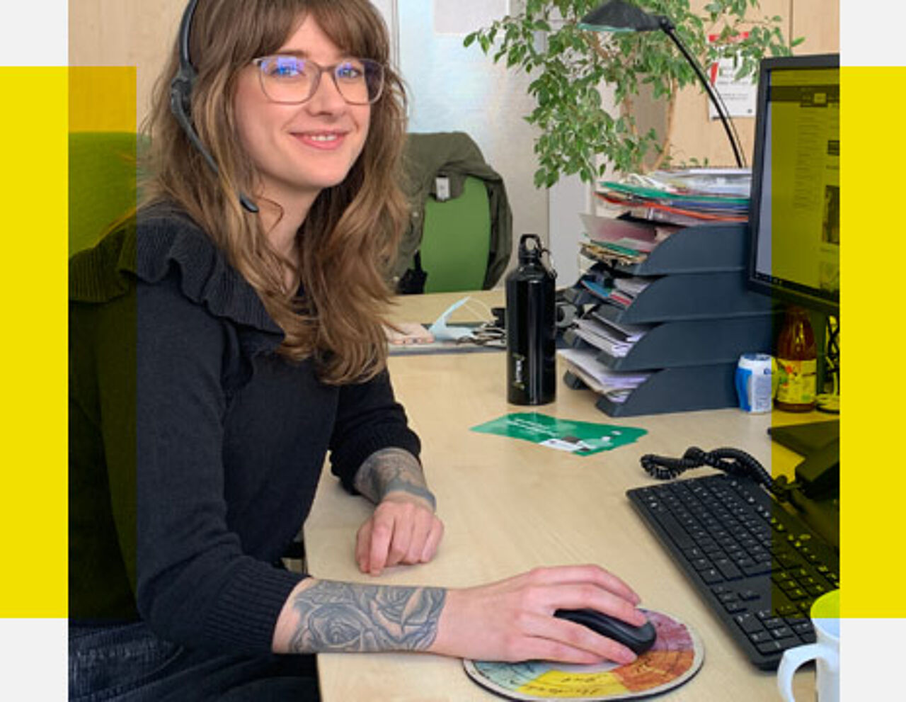 Campusspezialistin Lina sitzt mit Kopfhörern am Schreibtisch vor einem Bildschirm und schaut in Richtung Kamera