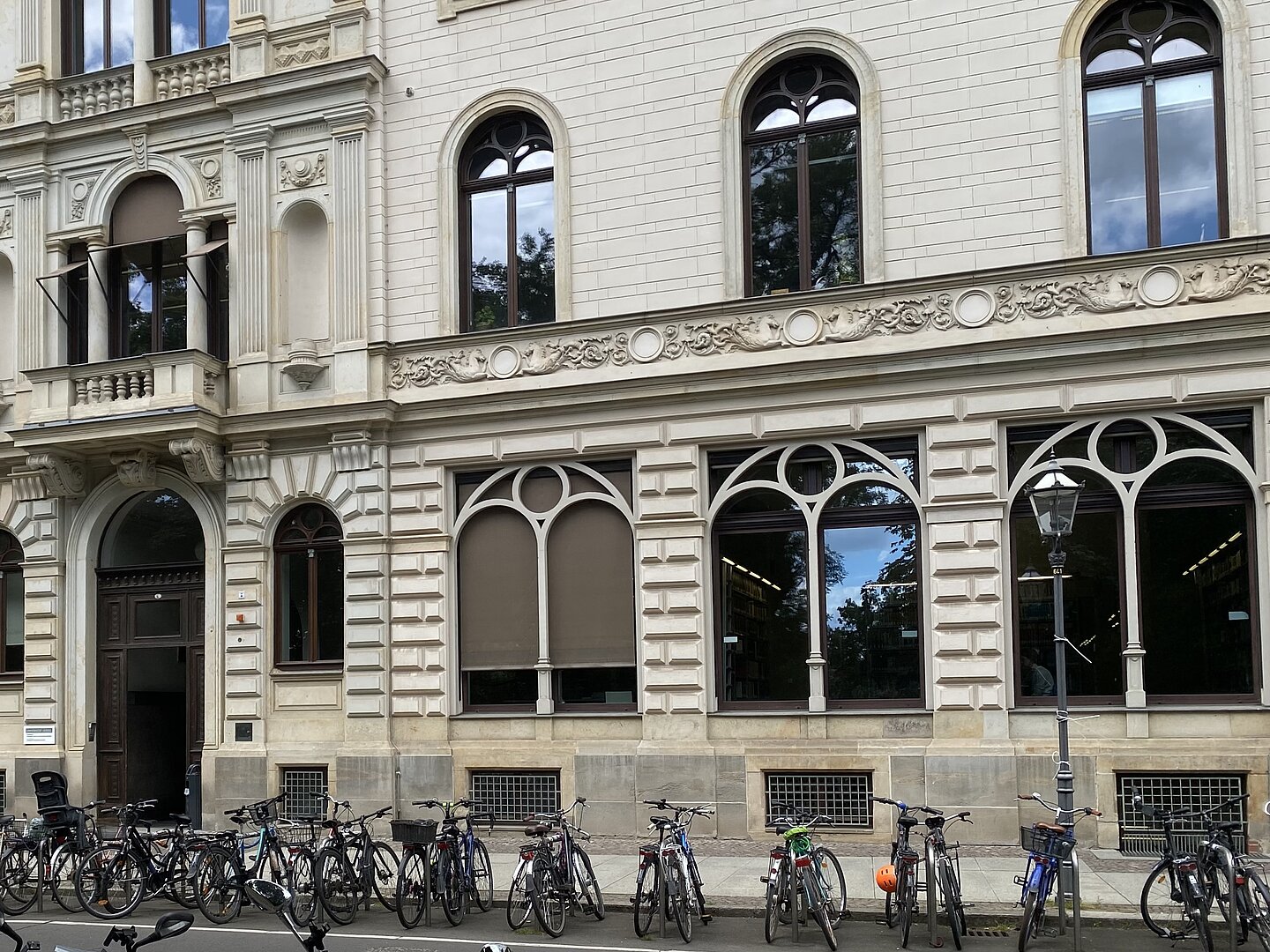 Ein altes Gebäude mit hell-greauen Fassaden. Die Fenster sind groß und gewolbt. Vor dem Gebäude stehen Fahrräder. Die große Eingangstür ist ebenfals gewölbt und besteht aus Holz.