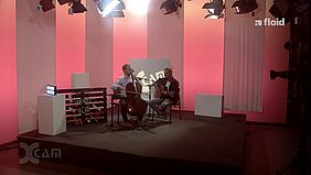 Screenshot eines Filmbeitrags, zwei Musiker mit Cello und Gitarre auf der Studiobühne