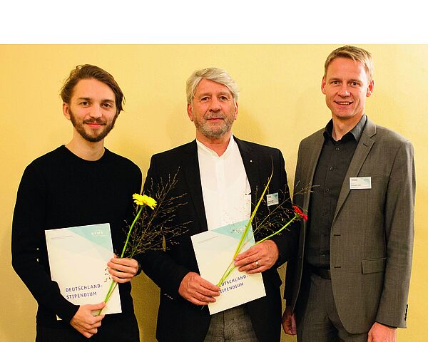 Drei Männer stehen vor einer gelben Wand un schauen in die Kamera. Zwei von ihnen haben einen Umschlag und Blumen in der Hand. Der dritte steht rechts neben ihnen. Sie tragen Anzüge.