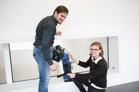 Zwei Studierende lächeln in die Kamera. Ein Student trägt ein intelligentes Bewegungsunterstützungssystem am Knie. Der andere Student kniet und hält die Kabel am Unterstützungssystem fest.