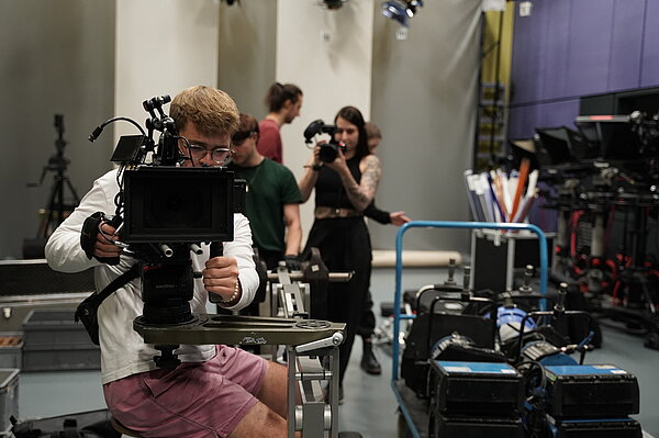 ein Mensch sitzt in Filmstudio hinter einer Kamera, die er bedient, im HIntergrund weitere Personen