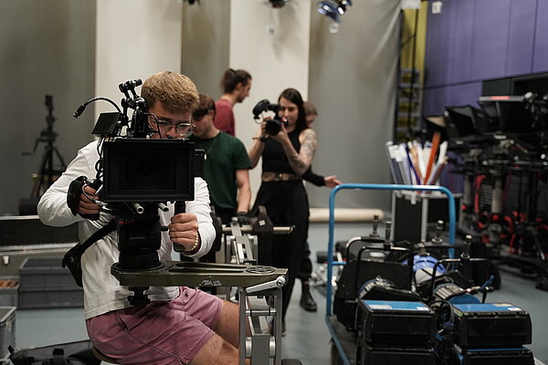 ein Mensch sitzt in Filmstudio hinter einer Kamera, die er bedient, im HIntergrund weitere Personen