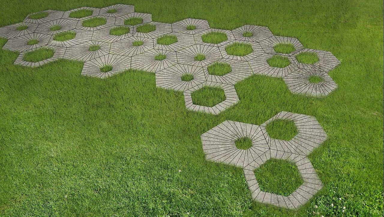 Betonpflaster-Kombination aus sechseckigen Steinen auf einer Rasenfläche. In den Steinen sind verschieden große Löcher, durch die das Gras wächst.
