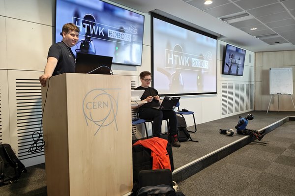 Vortrag der HTWK Robots im CERN (v.l.n.r. David Schulte, Max Polter, Ganymed) (c) HTWK Robots Person steht beim Vortrag am Pult vor einer Wand mit drei Monitoren. Daneben sitzt eine weitere Person mit Laptop auf den Knien und ein humanoider Roboter.