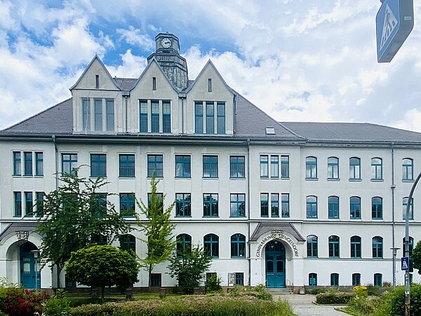Großes Gebäude mit weißer Fassade und grünen Türen und Fensterrahmen. Mittig auf dem Dach befindet sich ein kleiner sechseckiger Turm mit einer Uhr hinter 3 Dachgiebeln. Vor dem Gebäude befindet sich einige Beete.