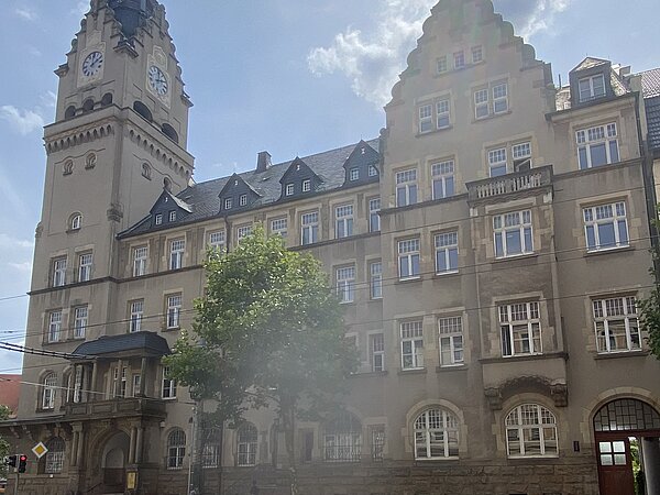 Ein alt verziertes Gebäude mit grau-brauner Fassade und anthrazitfarbenem Dach. Einige der Fenster haben einen Rundbogen. Vor dem Gebäude steht ein großer Baum.