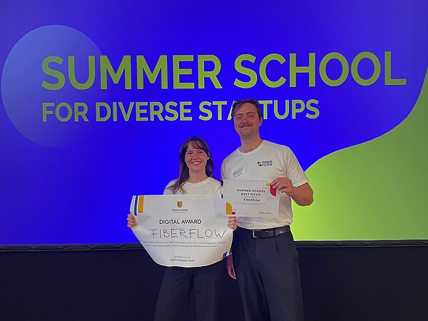 Lena und Ludwig von FiberFlow mit dem Gewinnercheck. Im Hintergrund große Leinwand mit der Aufschrift "Summer School for diverse Startups"