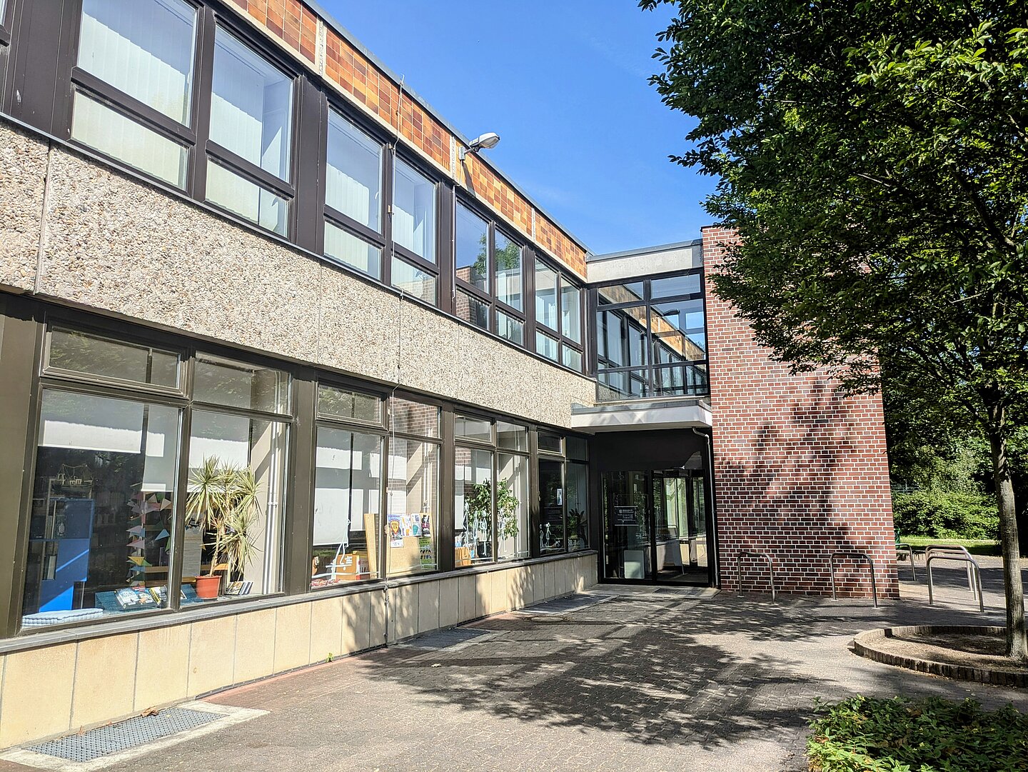 SB_Grünau_Süd.jpg Das Bild zeigt die Stadtbibliothek Grünau-Süd in Leipzig. Das Gebäude hat zwei Stockwerke und eine Fassade, die teils aus braunen Ziegelsteinen und teils aus hellgrauem Beton besteht. Die Fensterfronten sind groß und lassen viel Licht in das Innere des Gebäudes. Vor dem Eingang sind einige Fahrradbügel zu sehen. Im Vordergrund rechts steht ein Baum, der Schatten auf den Gehweg wirft. Der Himmel ist klar und blau.
