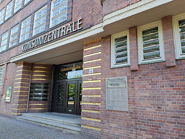 Der untere Teil eines roten Backsteingebäudes. Man sieht einige Fenster und zwei Treppenstufen, die zum Eingang führen, über dem in Großbuchstaben „Konsumzentrale“ steht. Der Eingang besteht aus schwarzen Türen und ist an der Fassade von goldfarbenen Akzenten umgeben.