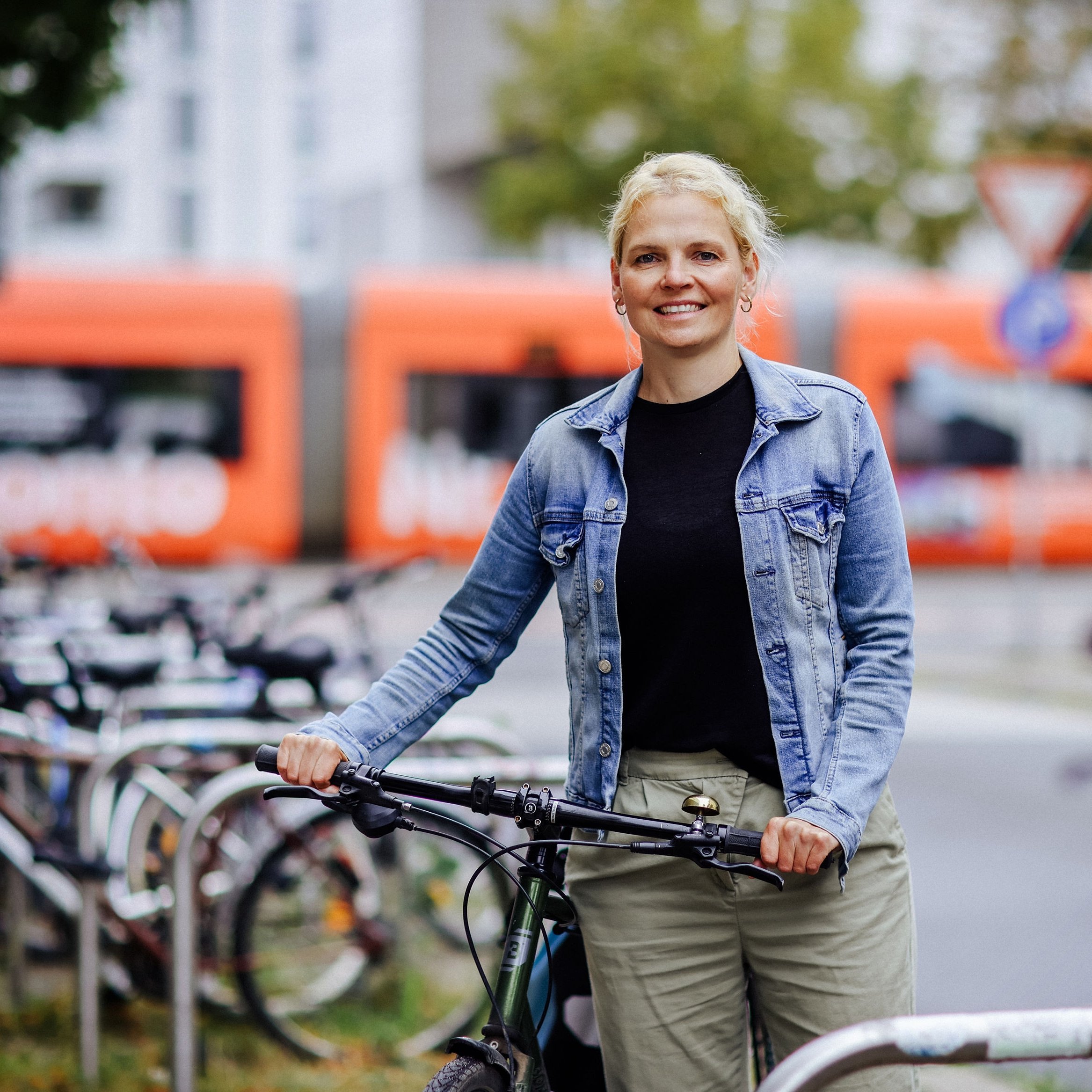 Professorin Ina Fichtner Lächelnde Frau an Fahrradständern auf dem HTWK Campus, im Hintergrund eine vorbeifahrende Straßenbahn.