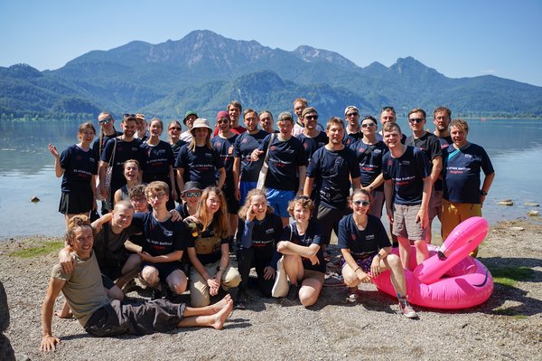 Fröhliches Gruppenbild der Teilnehmenden der Radtour im Teamshirt vor einem See mit Alpenpanorama.