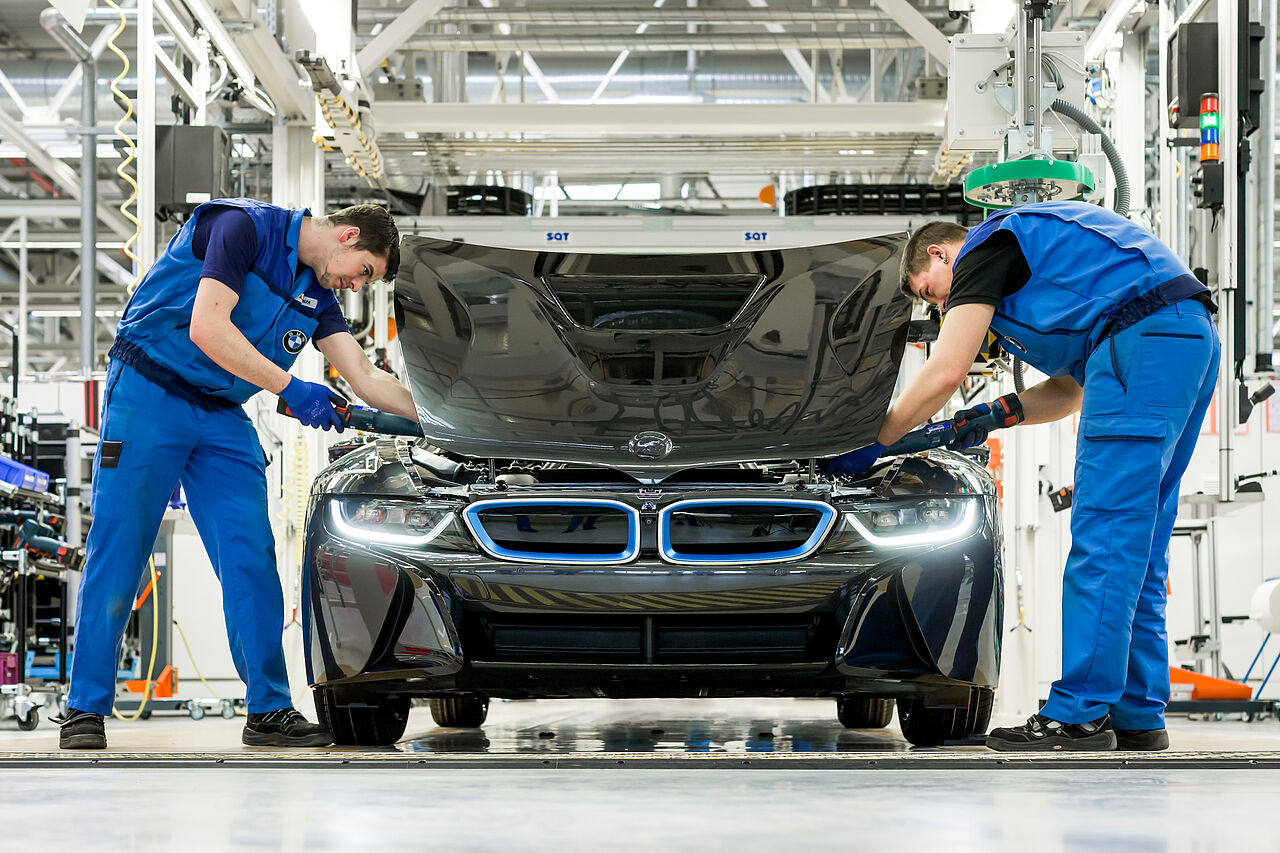Zwei Mitarbeiter des Leipziger BMW-Werks montieren die Motorhaube an einen Sportwagen. Dabei nutzt der linke Mitarbeiter einen etwas ergonomischeren Bewegungsablauf als der rechte 