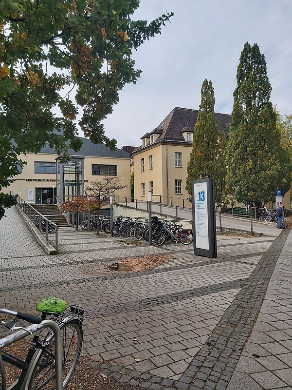 Das Bild zeigt eine ruhige Außenansicht eines modernen Gebäudekomplexes. Im linken Teil des Bildes ist ein Schild über dem Eingang zu erkennen, auf dem "Zentrum für Psychische Gesundheit" steht. Vor dem Eingang befindet sich eine Rampe, die für barrierefreien Zugang sorgt, sowie Treppen, die zum Haupteingang führen.