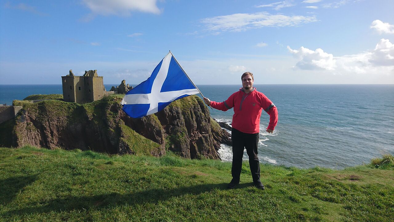 Aron Schaub schwenkt oben am Hang vor einer Steilküste am Meer die Schottlandfahne. Im Hintergrund die Überreste einer Burganlage.