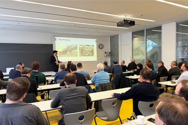 Foto von Seminarraum von hinten nach vorn aufgenommen, Teilnehmende sitzen an Tischen und hören Dozent zu, der beim Beamer-Bild steht und erklärt