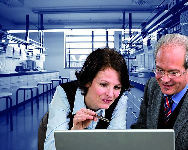 Eine Wissenschaftlerin und ein Wissenschaftler sitzen vor einem Laptop und besprechen ein Projekt. Im Hintergrund ist ein Labor zu sehen.