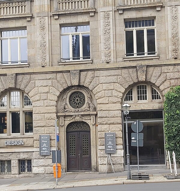 Das Bild zeigt die Fassade eines Gebäudes mit einer beeindruckenden, historischen Steinstruktur, die an klassische Architektur erinnert. Der Eingang ist kunstvoll mit einem runden Ornament über der Tür gestaltet, das ein auffälliges Detail darstellt. Um den Eingangsbereich herum befinden sich symmetrisch angeordnete Fenster mit kunstvollen Verzierungen. Die massiven Steine und die verzierten Elemente verleihen dem Gebäude einen repräsentativen und gleichzeitig robusten Charakter.