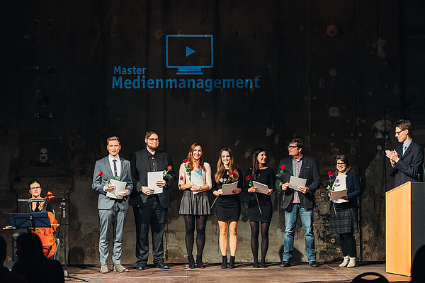 Eine Gruppe steht mit Rose und Mappe in der Hand auf der Bühne
