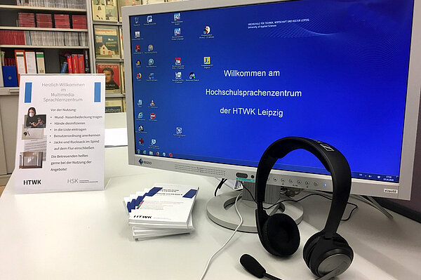 Monitor des Hochschulsprachenzentrums auf einem Schreibtisch mit Headset und Infomaterialien