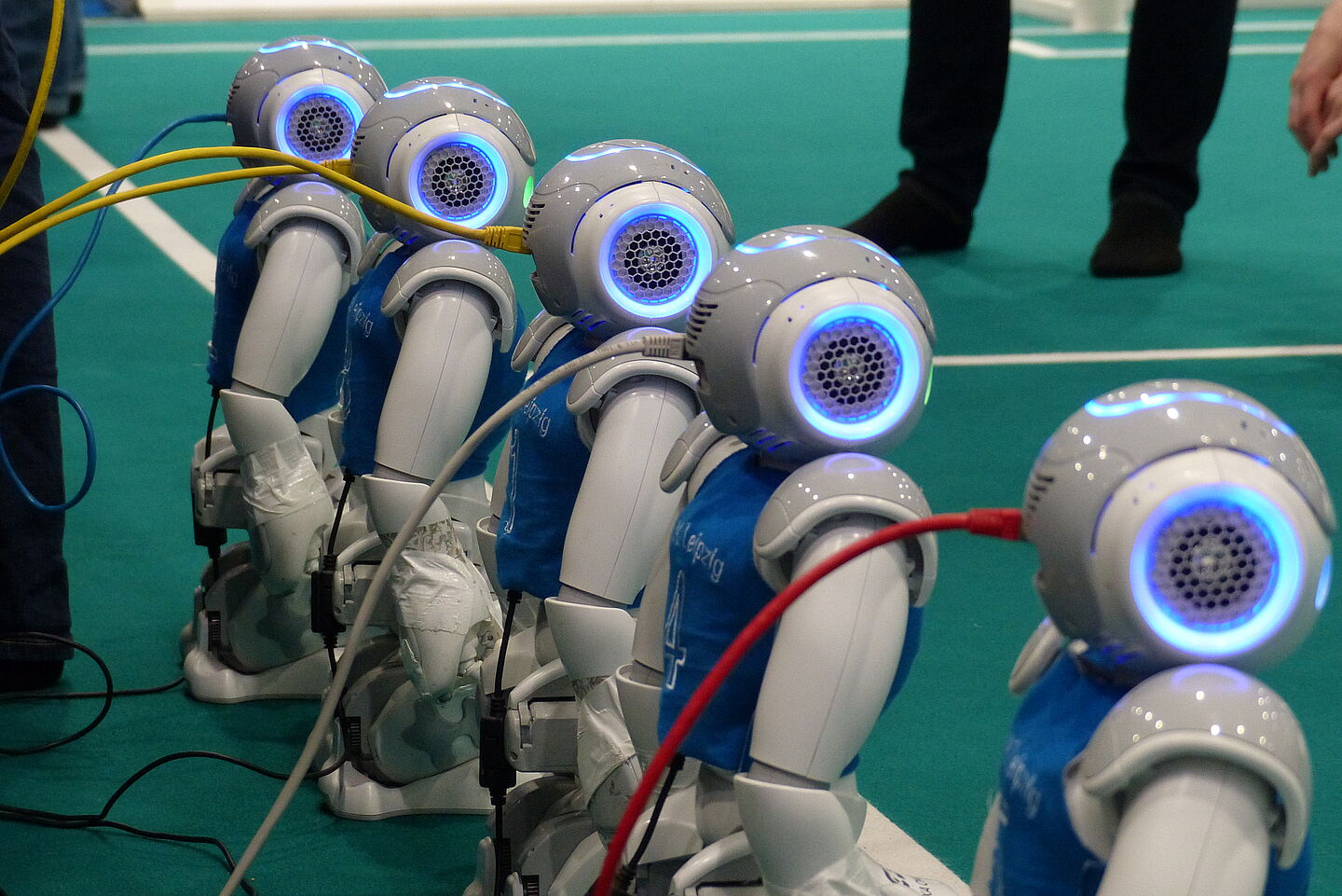 Nao-Robotermannschaft der HTWK Leipzig. (Foto: HTWK Leipzig) Fünf Nao-Roboter der HTWK Leipzig stehen nebeneinander am Spielfeldrand und sind mit Kabeln an einer Stromquelle angeschlossen.