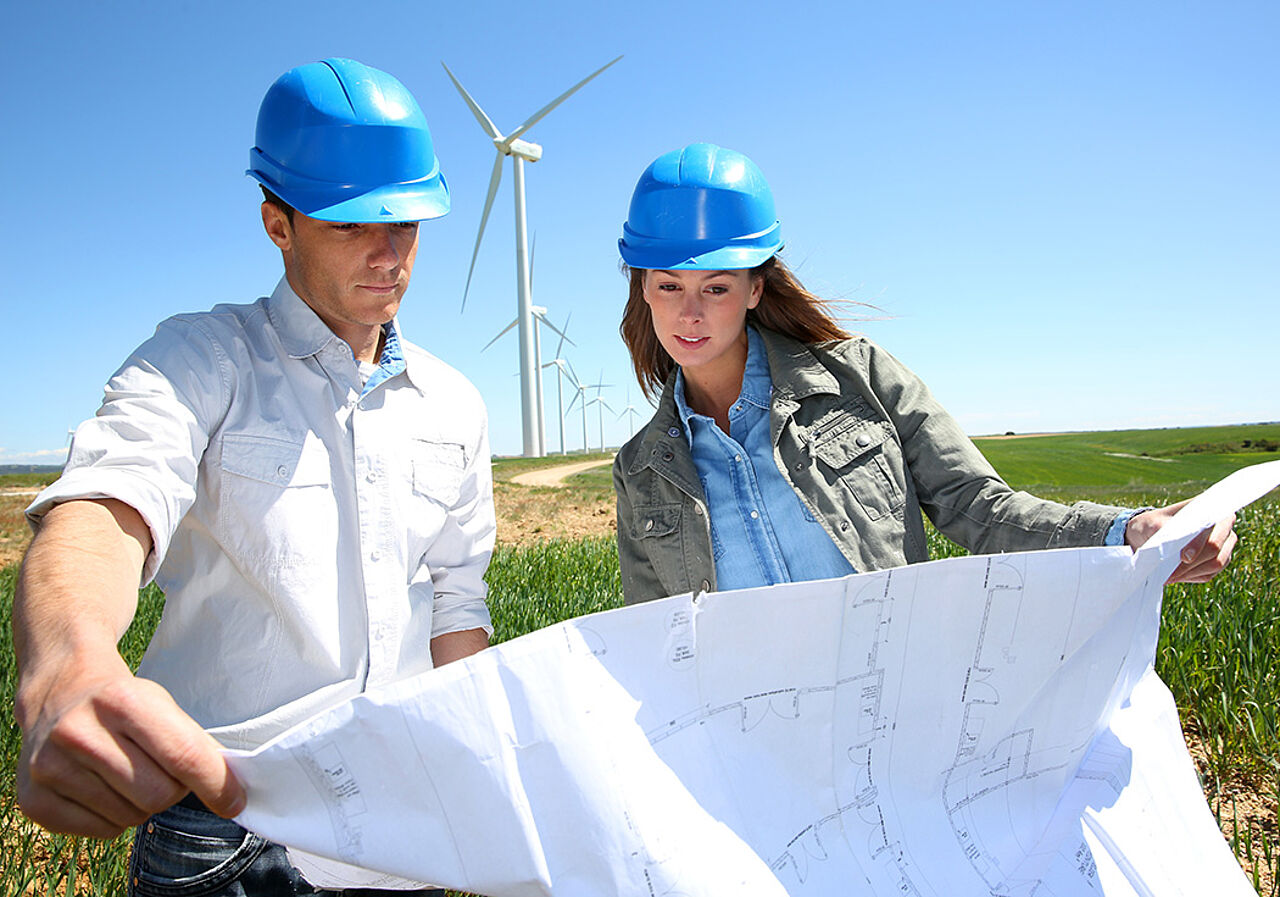 Eine weibliche Ingenieurin und ein männlicher Ingenieur betrachten einen Bauplan auf einem freien Feld mit Windrad