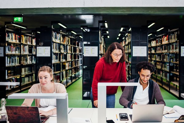 Eine Frau im roten Pulli gibt zwei Studierenden, die an ihren Notebooks in der Bibliothek arbeiten, Hinweise.