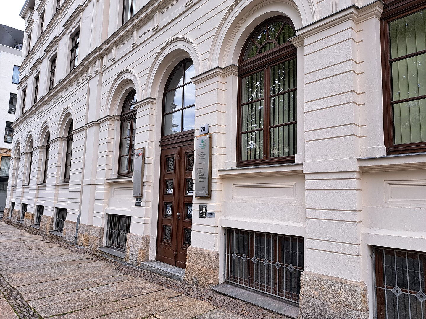 Das Bild zeigt das Leibniz-Institut für jüdische Geschichte und Kultur, ein historisches Gebäude mit klassischer Architektur. Die Fassade ist in Cremeweiß gehalten mit markanten, vertikalen Steinverzierungen und Fenstereinfassungen. Es verfügt über große, bogenförmige Fenster, die mit schmiedeeisernen Gittern geschützt sind. Eine massive Holztür mit einer Oberlichtverglasung dient als Eingang. Vor dem Gebäude erstreckt sich eine gepflasterte Straße. An der Tür ist eine Informationsplakette angebracht, die auf das Institut hinweist.