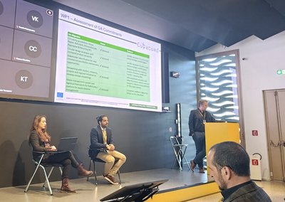 Prof. Böhm steht auf der Bühne und hält einen Vortrag. Neben ihm auf der Bühne sitzen zwei Menschen auf einem Stuhl