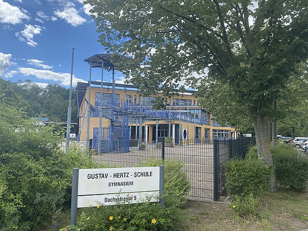 Gustav-Hertz-Gymnasium Ein asymmetrisches Gebäude mit gelber Fassade und blauem Geländer auf einem eingezäunten Gelände. Vor dem Zaun befindet sich ein Schild mit dem Namen der Schule. Das Gelände ist von vielen Bäumen umgeben.