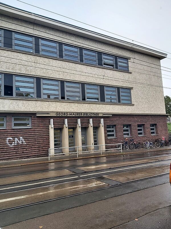 Das Bild zeigt die Fassade eines mehrstöckigen Gebäudes, das die „Georg-Maurer-Bibliothek“ beherbergt. Der Eingangsbereich befindet sich im unteren Teil des Gebäudes, welcher mit rötlich-braunen Ziegeln verkleidet ist, während die oberen Stockwerke mit einem helleren Putz versehen sind. Über dem Eingang steht der Name der Bibliothek in klaren Buchstaben.