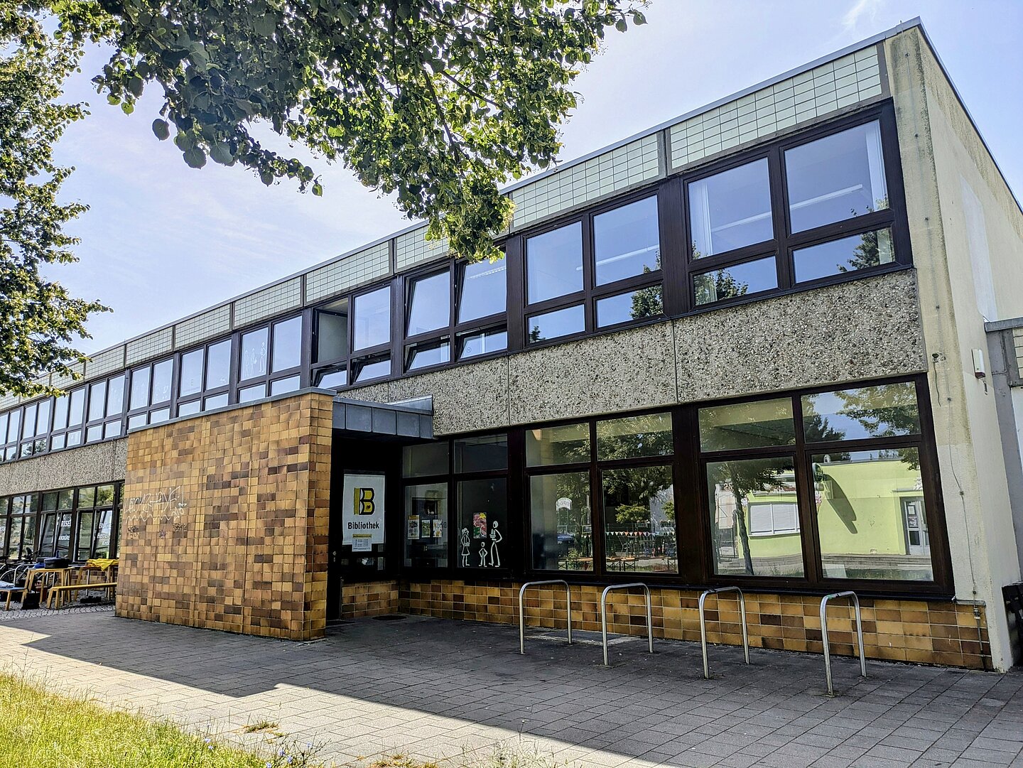 Die Stadtteilbibliothek Grünau Nord ist ein modernes, zweistöckiges Gebäude mit zahlreichen großen Fenstern. Der Eingang ist mit gelben Fliesen verkleidet und hat ein Schild mit der Aufschrift "Bibliothek". Vor dem Eingang befinden sich Fahrradständer. Im Vordergrund des Bildes sind Bäume und Sträucher zu erkennen