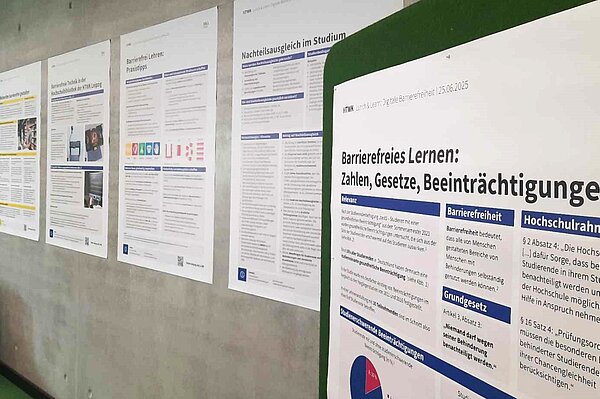 an einer grauen Betonwand sind Poster in Größe A0 in einer Reihe aufgehangen, im Vordergrund  ist das Poster mit dem Titel " Barrierefreies lernen: Zahlen, Gesetze, Beeinträchtigungen" zu sehen