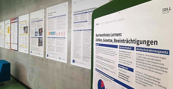 an einer grauen Betonwand sind Poster in Größe A0 in einer Reihe aufgehangen, im Vordergrund  ist das Poster mit dem Titel " Barrierefreies lernen: Zahlen, Gesetze, Beeinträchtigungen" zu sehen