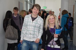 Zwei junge Teilnehmende lächeln in die Kamera, während sie in einem Flur mit Studierenden stehen