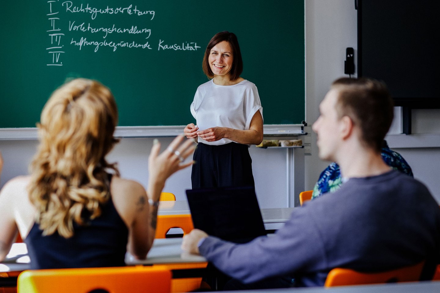 Dozentin vor Tafel mit Stichpunkten, Studierende diskutieren im Seminarraum.